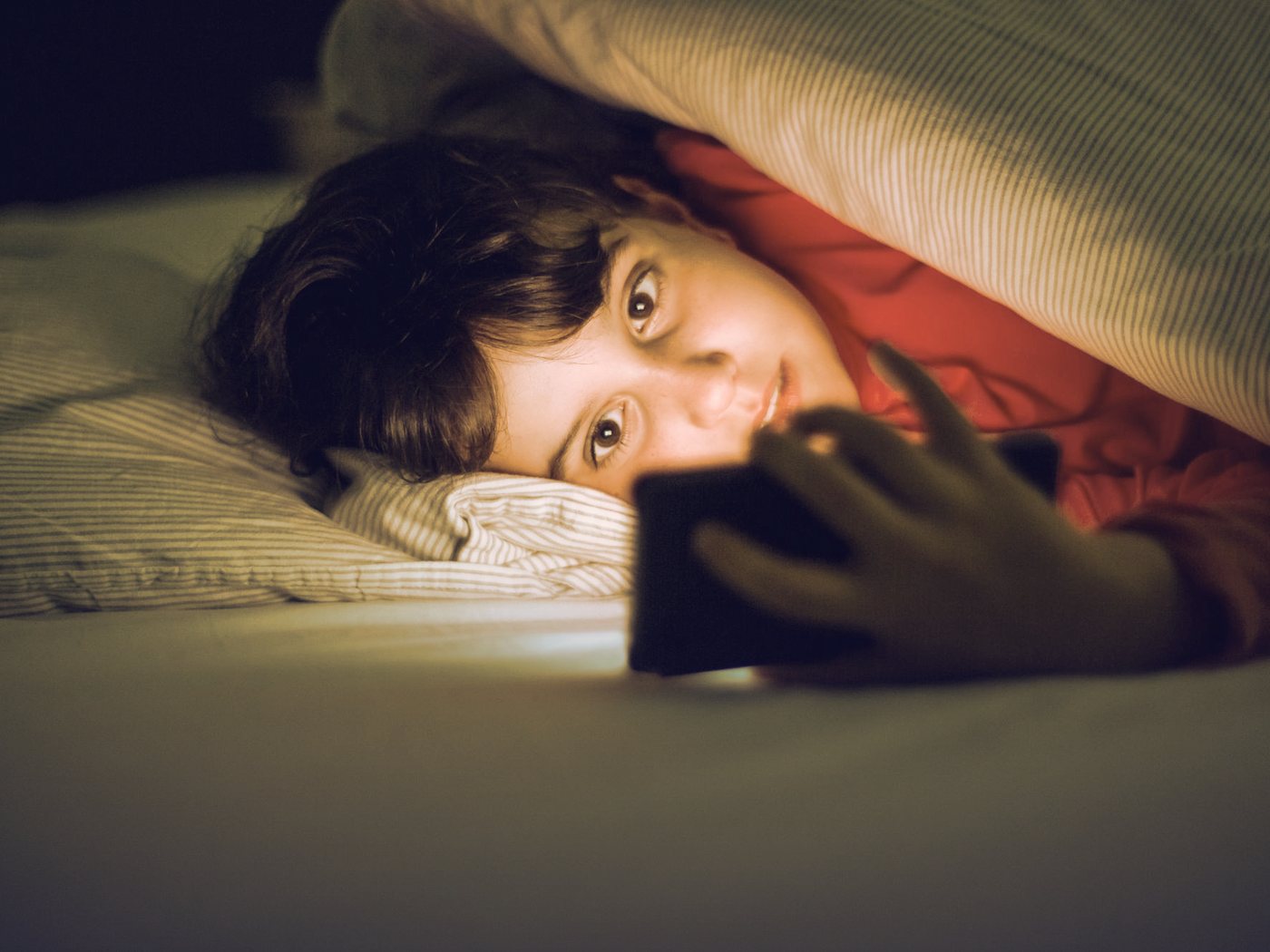 Estudiante bajo las sábanas mirando el celular de noche
