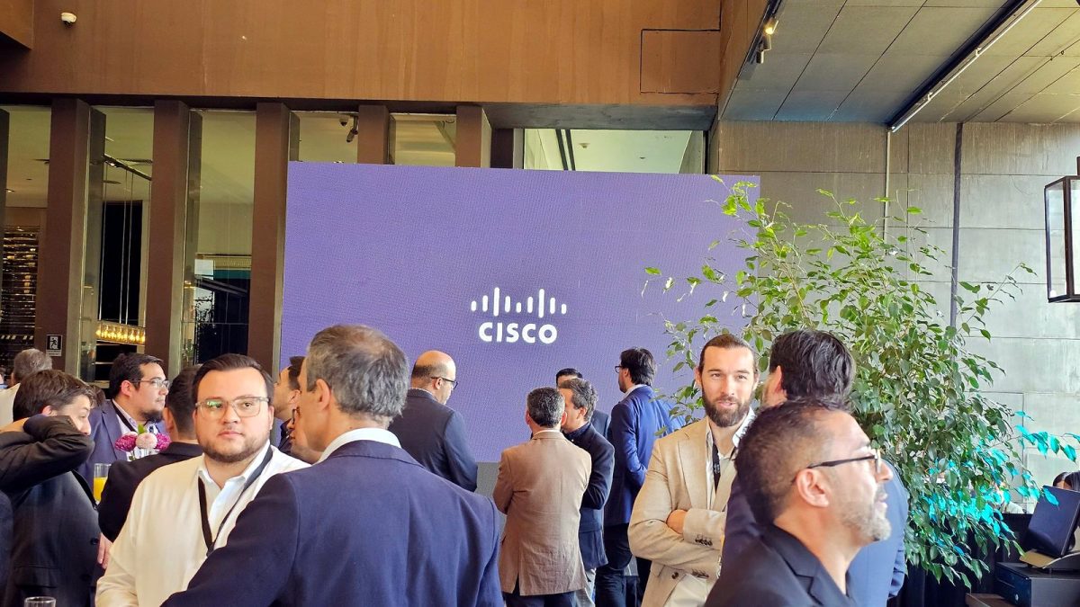Asistentes conversan en grupos frente a una pantalla con el logo de Cisco, junto a una planta.