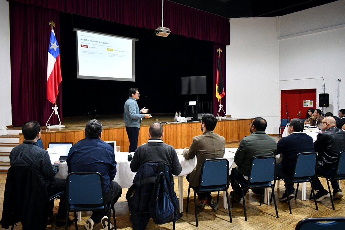 Un expositor presenta en el auditorio del Deutsche Schule de Viña del Mar, junto a las banderas de Chile y Alemania; en pantalla se lee «No todos los datos son iguales».